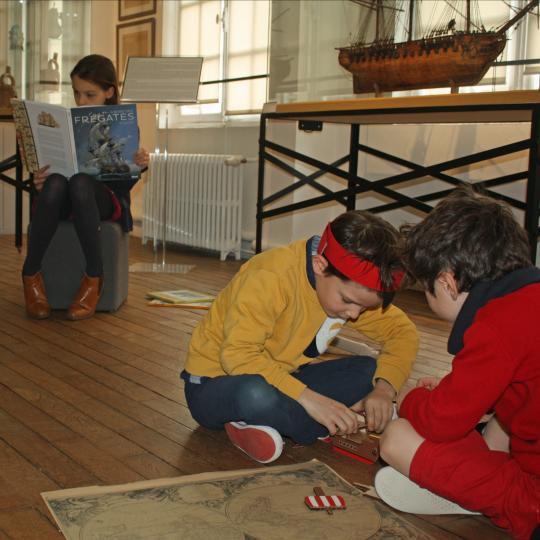 Trois enfants jouant avec le contenu de la malle corsaire: carte, maquette de bateau, boussole.