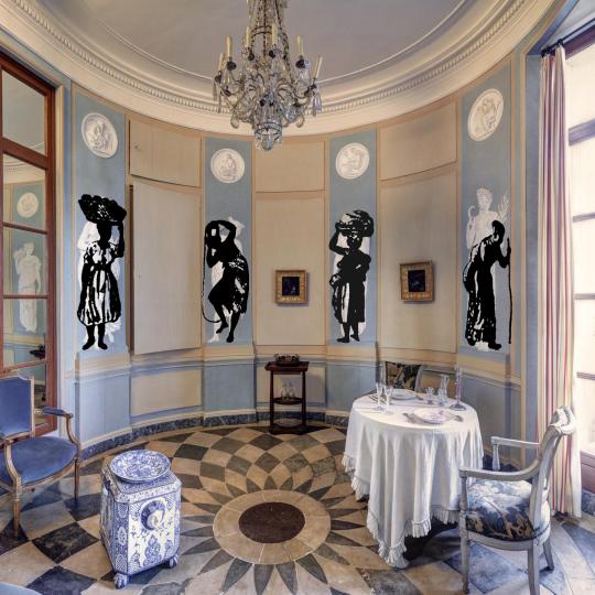 Photographie de la salle à manger de la Maison de l'armateur, avec la table dressée. Sur les murs, 4 projections de figures d'esclaves.