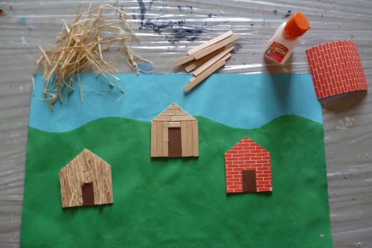 Maisonnettes en cartons, représentant trois types de matériaux (paille, bois, brique) collées dans un paysage.